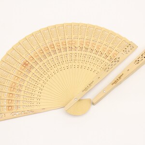 May include: A wooden hand fan with a delicate floral design and a matching handle. The fan is closed and the handle is engraved with the text "Natalie & Morgan, March 11, 2023".