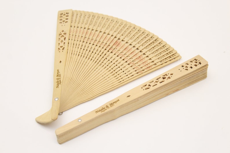 May include: Two wooden folding fans with intricate cutouts and personalised text. The text reads "Natalie & Morgan" and "August 12, 2023".