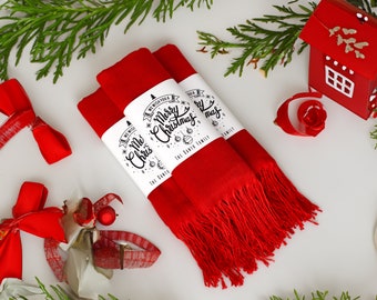May include: Three red scarves with fringe, each wrapped with a white band printed with "We Wish You a Merry Christmas" and "The Santa Family." The image includes red bows, a red house ornament, and greenery, creating a festive holiday scene.