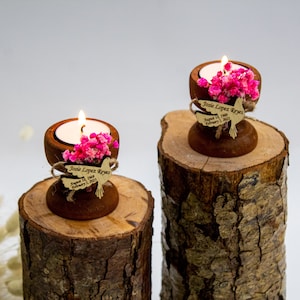May include: Two wooden candle holders with small tea lights, pink flowers, and personalized dove-shaped wooden tags. The tags read "Josie Lopez Reyes August 12, 1989 - February 1, 2023". The candle holders are sitting on top of two wooden logs.