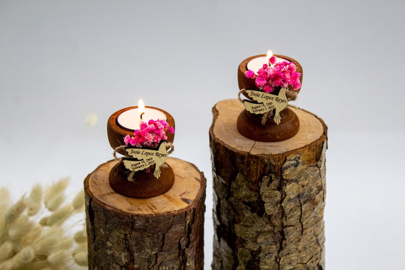 May include: Two wooden candle holders with small tea lights, pink flowers, and personalized dove-shaped wooden tags. The tags read "Josie Lopez Reyes August 12, 1989 - February 1, 2023". The candle holders are sitting on top of two wooden logs.