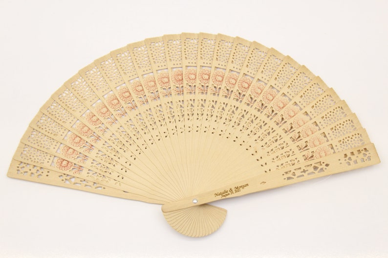 May include: A light brown wooden hand fan with a floral design and the text "Natalie & Morgan June 2021" engraved on the handle.