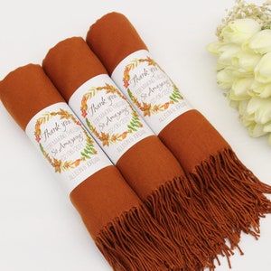 May include: Three rust-colored scarves with fringed edges, each wrapped with a white label that reads "Thank you for making our wedding so amazing!" with a floral design and the date "02/08/2021" and the name "Allison Jordan".