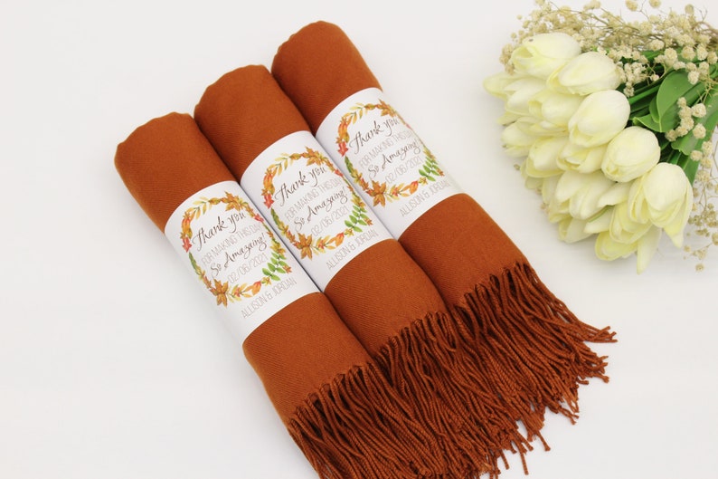 May include: Three rust-colored scarves with fringed edges, each wrapped with a white label that reads "Thank you for making our wedding so amazing!" with a floral design and the date "02/08/2021" and the name "Allison Jordan".