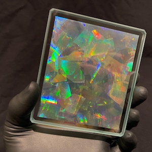 May include: A rectangular glass display piece featuring a multi-faceted holographic surface that reflects vibrant rainbow colors in geometric patterns. The clear glass frame has beveled edges, creating a prismatic effect across the entire surface area.