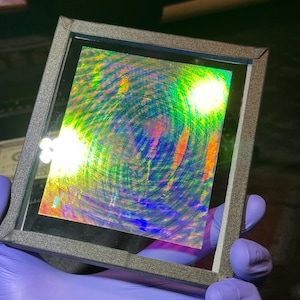 May include: A square, dark-framed display case containing a flat, iridescent sheet. The surface of the sheet features a vibrant, multi-colored holographic pattern with swirling, circular textures that shift in light, displaying hues of blue, green, and orange.