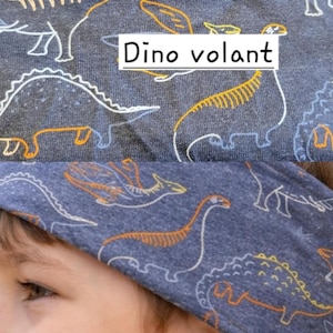 Peut inclure: Un bandeau bleu avec un motif de dinosaures blancs et orange. Le texte "Dino volant" est dans un encadré blanc. Le bandeau est porté par un enfant.