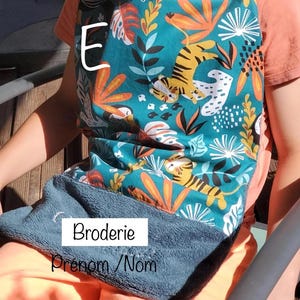 May include: A teal blue bib with a jungle print featuring orange tigers, green leaves, and white flowers. The bib has a gray pocket with the text "Broderie" and "Prenom/Nom". The bib is size XXL.