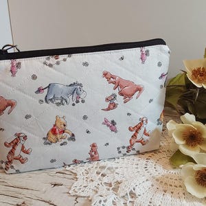 May include: A quilted fabric zip-up pouch featuring a repeating pattern of Winnie the Pooh characters including Eeyore, Tigger, Piglet, and Kanga on a light background. The pouch has a black zip closure along the top edge and a diamond-stitched texture.