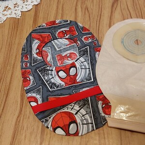May include: A gray and red fabric pouch with a Spiderman pattern and a red ribbon. The pouch is next to a beige ostomy bag with a clear, round, adhesive wafer.