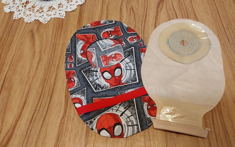 May include: A gray and red fabric pouch with a Spiderman pattern and a red ribbon. The pouch is next to a beige ostomy bag with a clear, round, adhesive wafer.
