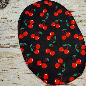 May include: Black oval-shaped fabric with a repeating pattern of red cherries and green leaves. The cherries are bright red, and the leaves are a vibrant green. The fabric is set against a rustic, weathered wooden background.