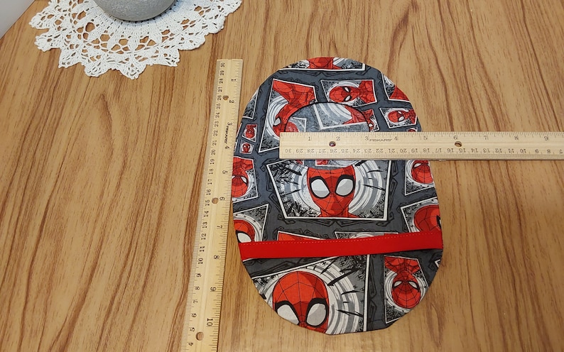 May include: A gray and red fabric circle with a repeating pattern of Spiderman's face in a comic book style. The fabric has a red trim around the edge. The circle is approximately 7 inches in diameter.