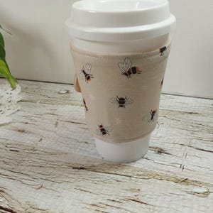 May include: A white travel mug with a beige fabric sleeve decorated with bee motifs. The mug has a white lid and sits on a wooden surface. The sleeve is fastened with a wooden button.