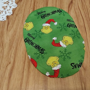 Peut inclure: Oval en tissu vert avec un motif répété du Grinch portant un chapeau de Père Noël et des flocons de neige. Le texte "Merry Grinchmas" est répété sur tout le motif.