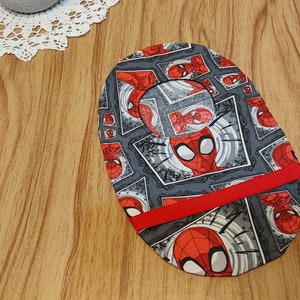 May include: A gray and red fabric oval with a repeated pattern of a cartoon superhero's face in a comic book frame. The fabric has a red ribbon sewn across the center.
