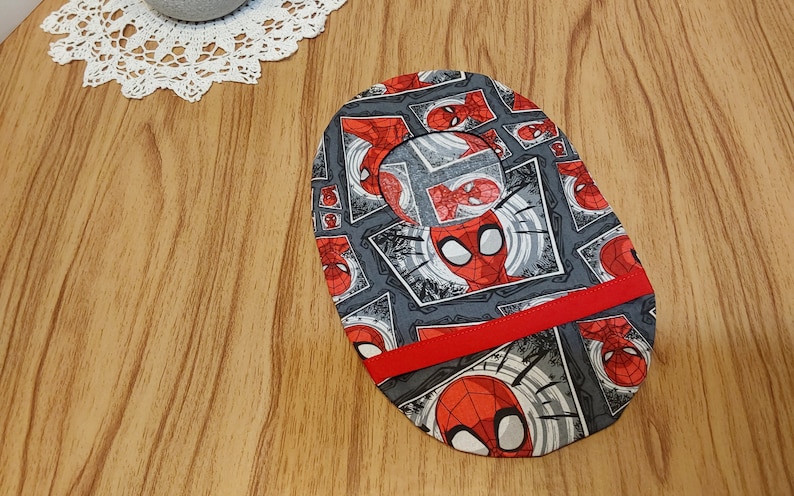 May include: A gray and red fabric oval with a repeated pattern of a cartoon superhero's face in a comic book frame. The fabric has a red ribbon sewn across the center.