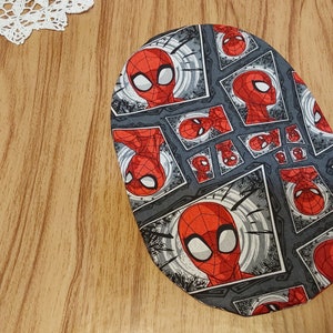 May include: A gray oval-shaped fabric with a repeating pattern of a red and black cartoon superhero face. The superhero is wearing a red and blue mask.