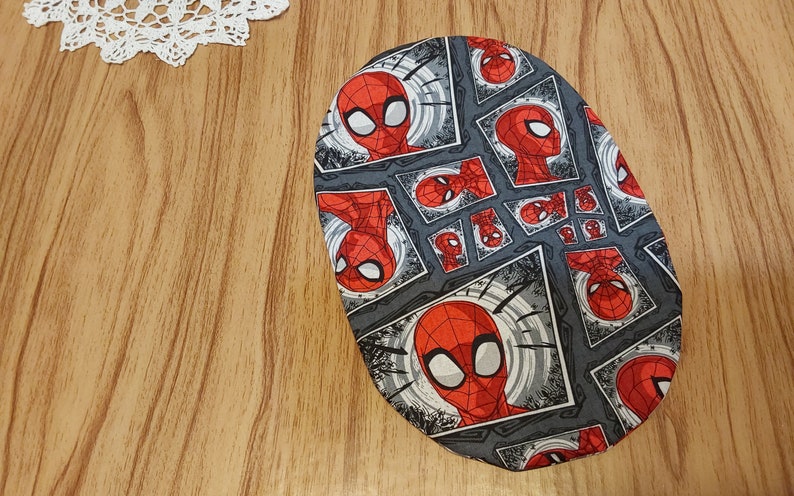 May include: A gray oval-shaped fabric with a repeating pattern of a red and black cartoon superhero face. The superhero is wearing a red and blue mask.
