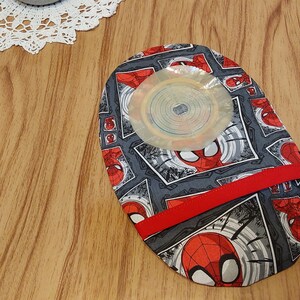 May include: A gray and red fabric pouch with a Spiderman pattern. The pouch has a red ribbon and a clear plastic circle in the center.