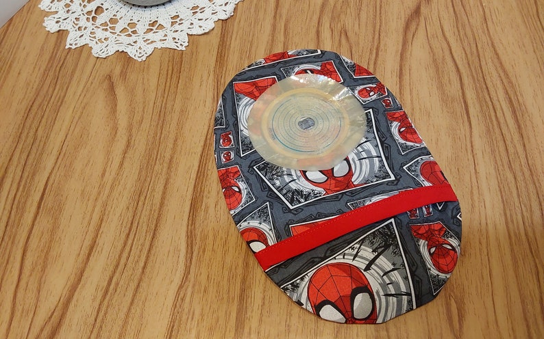 May include: A gray and red fabric pouch with a Spiderman pattern. The pouch has a red ribbon and a clear plastic circle in the center.