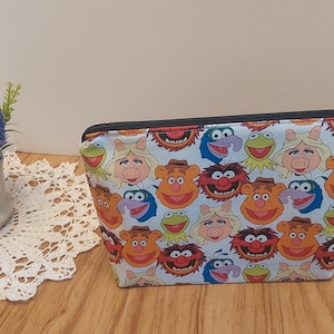 May include: A white zippered pouch with a colorful print of various Muppet characters, including Miss Piggy, Kermit the Frog, and Animal. The pouch has a black zipper and a silver ring.