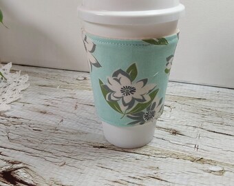 May include: A light blue fabric coffee cup sleeve with a floral pattern in white, gray, and green. The sleeve is wrapped around a white disposable coffee cup. The background is a light wood surface.