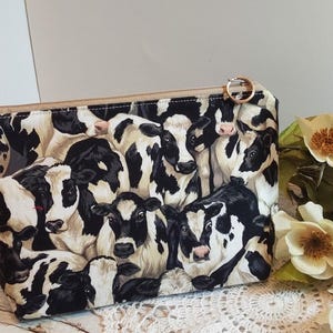 May include: A rectangular fabric pouch featuring a repeating black and white cow print pattern. The pouch has a tan zip closure along the top edge with a small circular metal ring attached to the zip pull. The fabric displays multiple cows.
