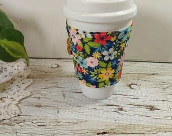 May include: A white ceramic travel mug with a floral patterned sleeve. The sleeve features a dark blue background with colorful flowers in shades of red, pink, yellow, and white. A wooden button secures the sleeve. The mug is on a distressed white wooden surface.
