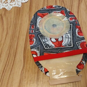 May include: A gray and red fabric pouch with a Spiderman design. The pouch has a clear plastic window and a red elastic band. The pouch is on a wooden surface.