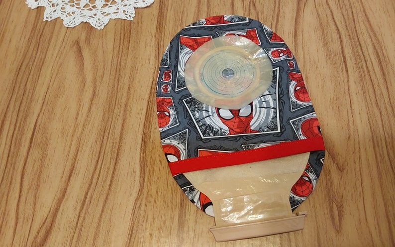 May include: A gray and red fabric pouch with a Spiderman design. The pouch has a clear plastic window and a red elastic band. The pouch is on a wooden surface.