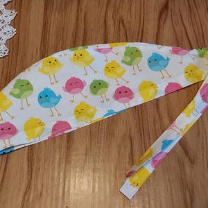 May include: A white scrub cap with a colorful pattern of cartoon birds. The birds are yellow, pink, blue, and green. The cap has a long tie that can be tied at the back of the head.