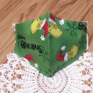 May include: Green fabric face mask with a repeating pattern of the Grinch wearing a Santa hat and snowflakes. The text "Merry Grinchmas" is printed on the mask.