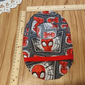 May include: A gray and red fabric baby bib with a Spiderman pattern. The bib has a red trim around the edges.