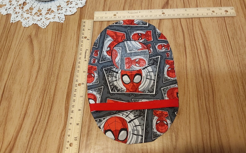 May include: A gray and red fabric baby bib with a Spiderman pattern. The bib has a red trim around the edges.