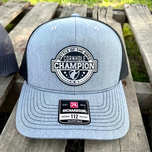 Pode incluir: Um chapéu de caminhoneiro cinza e preto com um remendo branco que diz "Battle of the Bags Cornhole Champion" com um gráfico de saco de cornhole. O chapéu é ajustável e tem o logotipo da Richardson e o texto "Original 112 Trucker".