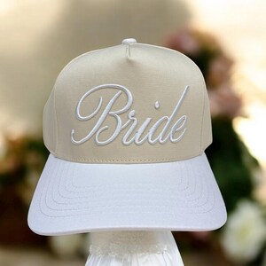 May include: A beige and white baseball cap featuring the word "Bride" embroidered in white cursive lettering. The cap has a flat peak and a structured crown, suitable for a wedding or hen party.