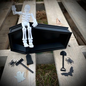 May include: A white skeleton figurine sitting in a black coffin with a black shovel, rake, tombstone, spider, and bat beside it.