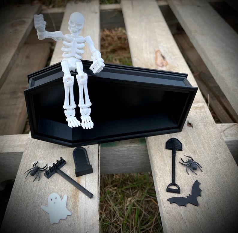 May include: A white skeleton figurine sitting in a black coffin with a black shovel, rake, tombstone, spider, and bat beside it.