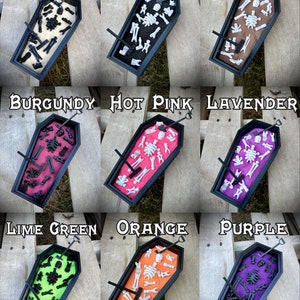 May include: Nine miniature coffins filled with sand and tiny skeleton bones. Each coffin is a different color: beige, black, brown, burgundy, hot pink, lavender, lime green, orange, and purple.