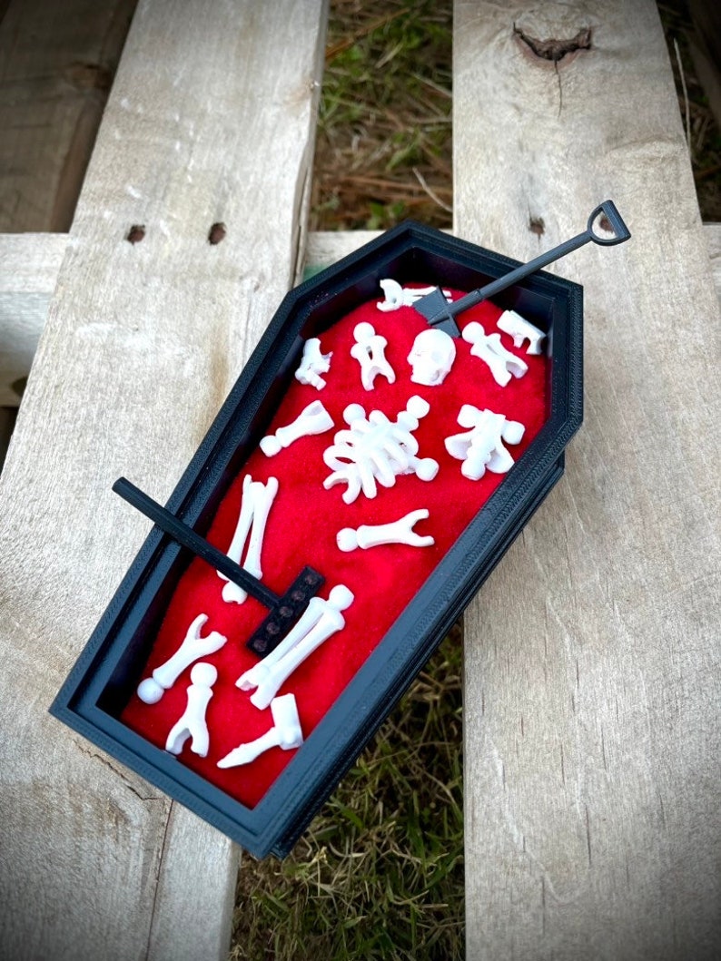 May include: A black coffin-shaped box with a red velvet interior. The box is filled with small white plastic bones and a tiny shovel. The box is sitting on a wooden surface.