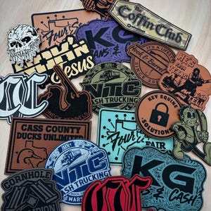 Peut inclure: Une collection d'écussons de formes variées présentant des motifs divers, notamment des crânes, des camions, des contours d'États et des textes comme The Coffin Club, Cass County Ducks Unlimited, NTC Nash Trucking et Cornhole Champion.