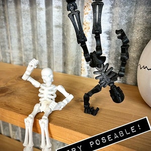May include: Two articulated skeleton figures, one black and one white, are posed on a wooden shelf. The black skeleton is doing a handstand, while the white skeleton is sitting with one arm raised. The text "VERY POSEABLE!" is printed on a black label on the shelf.