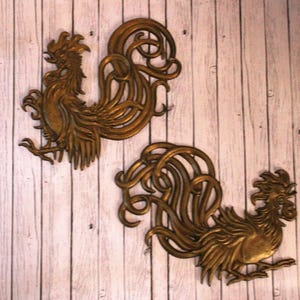 May include: Two bronze-colored rooster wall sculptures. Each rooster is detailed with a textured body and elaborate tail feathers. The sculptures are mounted on a light-colored, vertical-grain wooden surface.