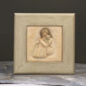 May include: A framed, carved wooden figurine depicts a person embracing a cat. The figure is in a light-colored dress, and the frame is a muted green. The artwork is displayed in a square format.