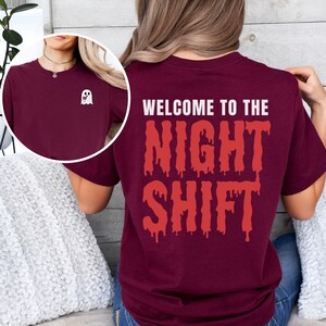 Peut inclure: Un t-shirt bordeaux avec le texte "Welcome to the Night Shift" en lettres rouges qui coulent. Un petit fant&ocirc;me blanc avec un c&oelig;ur rouge est sur le devant du t-shirt.