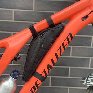 May include: An orange Specialized mountain bike frame with a black storage bag attached. The bag is secured with black straps. A water bottle is visible near the frame. The bike is against a brick wall.