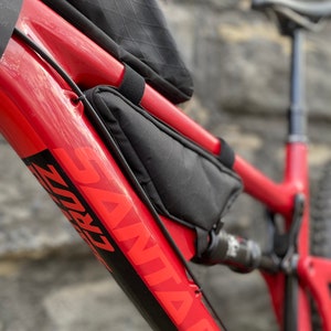 May include: Close-up of a red mountain bike frame with a black and red "SANTA CRUZ" logo. The bike has a black frame bag attached to the top tube.