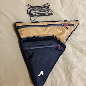 May include: A black and tan triangular bike bag with a zipped pocket and a white logo. The bag is on a beige surface with a black cord.