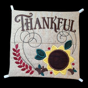 May include: A beige fabric tissue box cover with a sunflower, leaves, and acorns embroidered on it. The words "THANKFUL" are embroidered in brown lettering at the top.